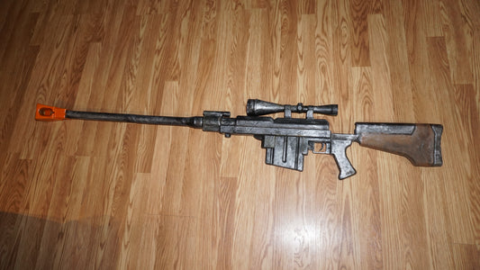 A large, black and silver toy gun with a scope and orange tip is lying on a wooden floor.